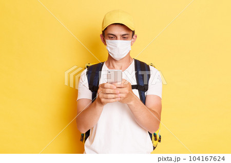 indoor shot of delivery man in yellow cap, t-shirt and protective mask, with thermal food backpack using cell phone while standing isolated on yellow background. indoor shot of delivery man in yellow cap, t-shirt and protective mask, with thermal food backpack using cell phone while standing isolated on yellow background. 104167624