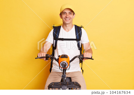 Food delivery. Portrait of smiling positive courier guy riding bike with backpack bag delivering meal from takeaway cafe or restaurant isolated over yellow background, 104167634