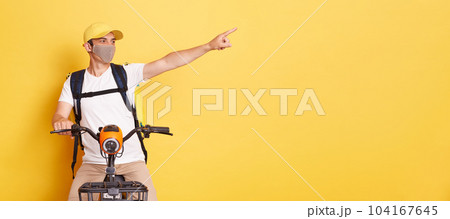 Horizontal shot of smiling positive delivery man in protective mask with thermo backpack, pointing at advertisement area, delivering orders on electric scooter, isolated on yellow background. Horizontal shot of smiling positive delivery man in protective mask with thermo backpack, pointing at advertisement area, delivering orders on electric scooter, isolated on yellow background. 104167645