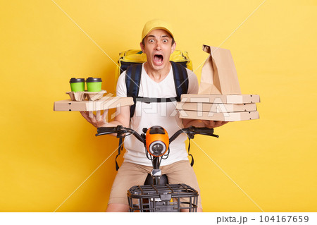 Shocked courier wearing cap and white t shirt holding pizza boxes and coffee, riding bicycle looking at camera with open mouth and big eyes, being scared, isolated on yellow background 104167659