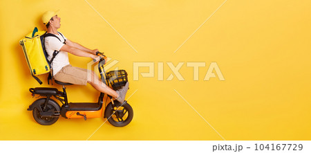 Full length side view portrait of man courier with thermo backpack in white T-shirt and cap riding electric bike, braking, avoids accident, looks scared and shocked, isolated over yellow background. 104167729