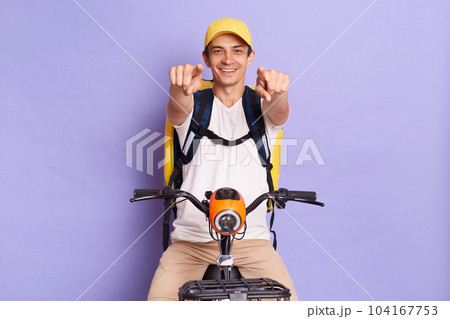 Indoor shot of young satisfied delivery man of takeaway food on electric posing isolated over purple background, pointing at you with happy facial expression. Indoor shot of young satisfied delivery man of takeaway food on electric posing isolated over purple background, pointing at you with happy facial expression. 104167753
