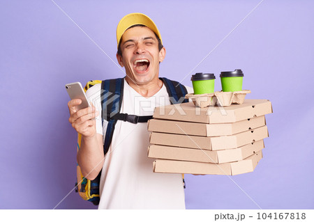 Extremely happy man courier in yellow cap and T-shirt holding food order pizza boxes and coffee to go, standing with phone in hands, screaming happily, isolated on purple background. Extremely happy man courier in yellow cap and T-shirt holding food order pizza boxes and coffee to go, standing with phone in hands, screaming happily, isolated on purple background. 104167818