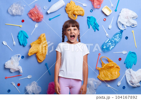 Angry desperate little girl wearing white t shirt posing against blue wall and much plastic garbage around, screaming loud, wants to be heard, shouting about danger for our planet. Angry desperate little girl wearing white t shirt posing against blue wall and much plastic garbage around, screaming loud, wants to be heard, shouting about danger for our planet. 104167892