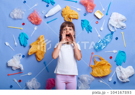Portrait of charming little girl with braids wearing white t shirt standing against much plastic wastes on blue wall, keeps hands near mouth, screaming about pollution on the Earth. Portrait of charming little girl with braids wearing white t shirt standing against much plastic wastes on blue wall, keeps hands near mouth, screaming about pollution on the Earth. 104167893