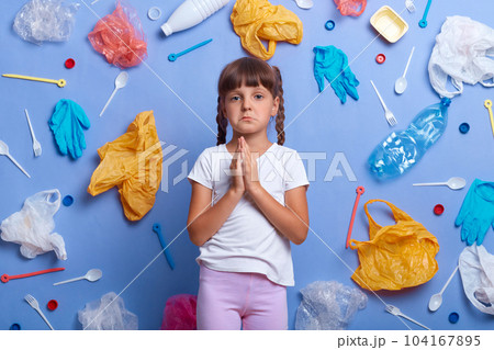 Sad crying little girl dressed casually standing with palms together, praying for Earth, looking at camera with pout lips, prays for ecosystem, poses against blue wall with much waste around. 104167895
