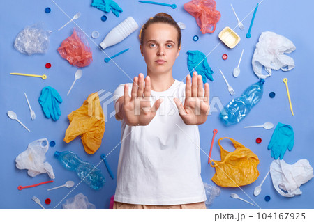 Portrait of serious Caucasian woman showing no gesture, demands to stop pollution, being eco friendly, poses against much plastic wastes against blue wall, angry with environmental contamination. Portrait of serious Caucasian woman showing no gesture, demands to stop pollution, being eco friendly, poses against much plastic wastes against blue wall, angry with environmental contamination. 104167925