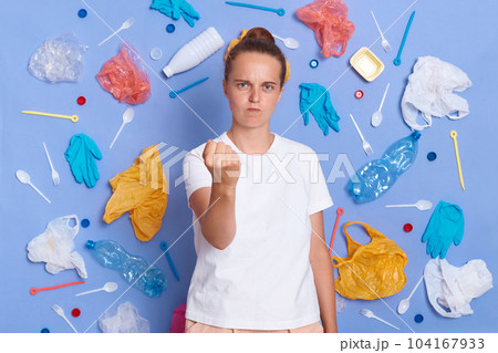 Image of angry aggressive woman dressed in white T-shirt surrounded with picked plastic litter isolated on blue wall, showing fist to camera, wants to punish those who harm nature. Image of angry aggressive woman dressed in white T-shirt surrounded with picked plastic litter isolated on blue wall, showing fist to camera, wants to punish those who harm nature. 104167933