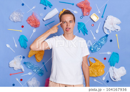 Portrait of funny woman dressed in white T-shirt surrounded with picked plastic litter isolated on blue wall, making suicide gesture and showing tongue out, being tired of pollution. 104167936