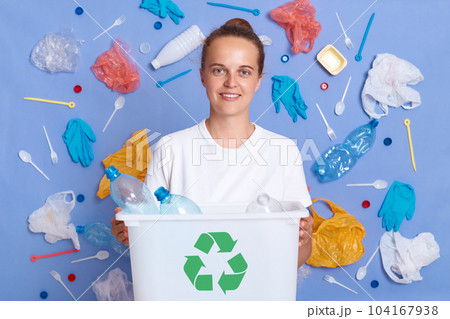 Portrait of happy responsible Caucasian woman eco activist with box full of plastic bottles for recycling, looking at camera, picked plastic litter isolated on blue wall. 104167938