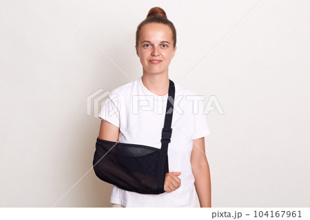 Smiling Caucasian woman broke arm during workout, take pain pills, wears arm sling, has good mood does not feel pain, wearing white t shirt, visit doctor, got into accident. Smiling Caucasian woman broke arm during workout, take pain pills, wears arm sling, has good mood does not feel pain, wearing white t shirt, visit doctor, got into accident. 104167961