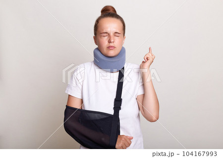 Indoor shot of hopeful woman with bun hairstyle wearing casual T-shirt and Shants collar, standing in arm sling, posing with closed eyes and crossed fingers, isolated on light gray background. Indoor shot of hopeful woman with bun hairstyle wearing casual T-shirt and Shants collar, standing in arm sling, posing with closed eyes and crossed fingers, isolated on light gray background. 104167993