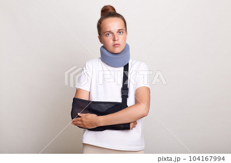 Image of sick injured woman with bun hairstyle wearing casual T-shirt and Shants collar, looking at camera, holding her hands, felling pain, standing in arm sling isolated on light gray background. 104167994
