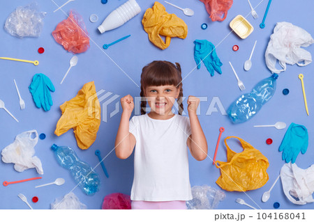 Angry aggressive little girl wearing white t shirt posing against blue wall and much plastic garbage around, clenched fists and screaming with hate, wants to save planet from plastic pollution. 104168041