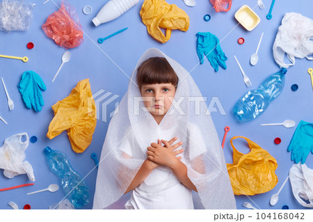 Portrait of serious upset little girl posing against blue background, surrounded with rubbish, looking at camera, saving planet from plastic pollution, has much thoughts, picks up litter for recycling 104168042