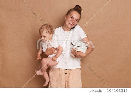 Indoor shot of cheerful joyful Caucasian mother with bun hairstyle holding her infant daughter and pot with porridge or soup, talking phone isolated over brown background. Indoor shot of cheerful joyful Caucasian mother with bun hairstyle holding her infant daughter and pot with porridge or soup, talking phone isolated over brown background. 104168315