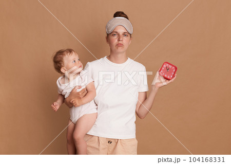 Indoor shot of sad upset Caucasian woman with dark hair wearing sleeping standing with her baby daughter in hands and showing alarm clock, sleepy mother needs more rest. Indoor shot of sad upset Caucasian woman with dark hair wearing sleeping standing with her baby daughter in hands and showing alarm clock, sleepy mother needs more rest. 104168331