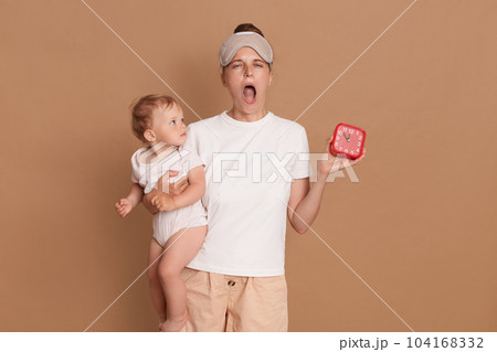 Portrait of tired sleepy woman wearing white t shirt and blindfold, yawning with widely open mouth, holding baby daughter and red alarm clock, time to wake up, posing isolated over brown background. 104168332