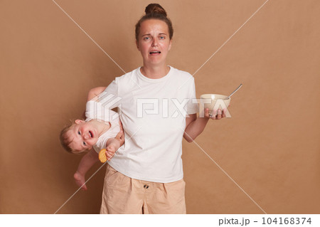 Portrait of shocked crying screaming woman wearing white T- shirt standing with her little daughter in hands isolated over brown background, trying to feed her child. Portrait of shocked crying screaming woman wearing white T- shirt standing with her little daughter in hands isolated over brown background, trying to feed her child. 104168374