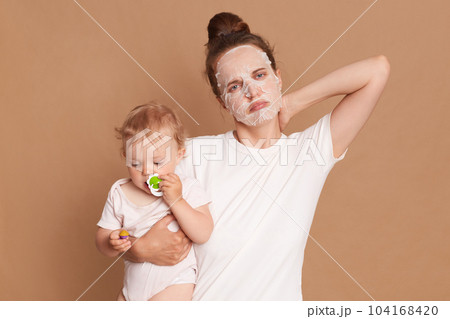 Indoor shot of sad unhealthy woman wearing white t shirt doing cosmetic procedures, feels unwell, has pain neck, looking at camera with sad expression, holding baby daughter in hands. Indoor shot of sad unhealthy woman wearing white t shirt doing cosmetic procedures, feels unwell, has pain neck, looking at camera with sad expression, holding baby daughter in hands. 104168420
