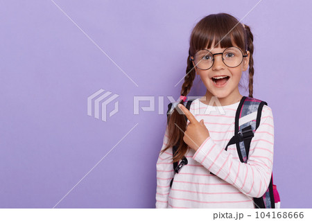 Horizontal shot of excited schoolgirl pointing with her fingers aside, showing copy space for promotional text, wearing striped shirt and glasses isolated on violet background. 104168666