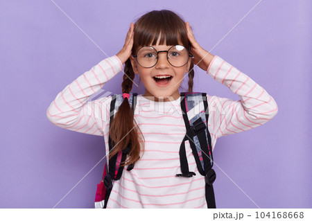 Portrait of excited amazed little girl with pigtails wearing backpack standing isolated over purple background, posing with raised arms, touching head, has great news. 104168668