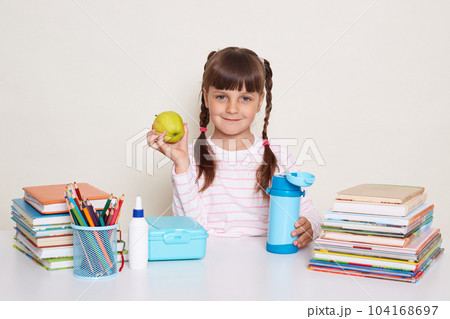 Horizontal shot of smiling optimistic little schoolgirl with pigtails wearing striped shirt sitting at the desk surrounded with books and other school supplies, holding fresh fruit, looking at camera. Horizontal shot of smiling optimistic little schoolgirl with pigtails wearing striped shirt sitting at the desk surrounded with books and other school supplies, holding fresh fruit, looking at camera. 104168697
