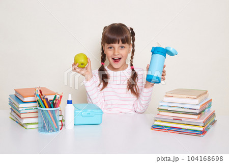 Portrait of satisfied amazed little girl with braids sitting at desk posing isolated over white background, being happy to have and snack during break, holding apple and bottle with water. Portrait of satisfied amazed little girl with braids sitting at desk posing isolated over white background, being happy to have and snack during break, holding apple and bottle with water. 104168698