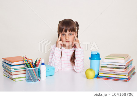 Image of sad serious little schoolgirl with pigtails wearing striped shirt, keeps fingers on temples, looking at camera, sitting at the desk surrounded with books and other school supplies. Image of sad serious little schoolgirl with pigtails wearing striped shirt, keeps fingers on temples, looking at camera, sitting at the desk surrounded with books and other school supplies. 104168706
