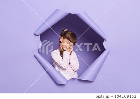 Portrait of sleepy tired adorable little girl with braids looking through breakthrough of purple background, standing with closed eyes, leaning on her palms, feels sleepless. 104169152