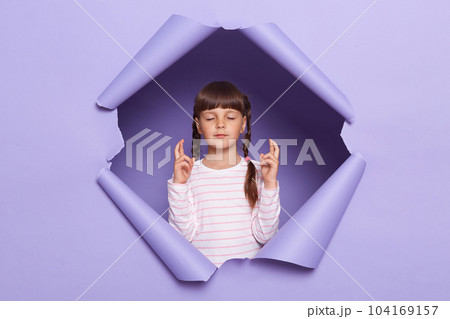 Image of charming little girl with braids wearing striped shirt posing in torn paper wall, standing with closed eyes and crossed fingers, making wish, dreaming of the best. 104169157