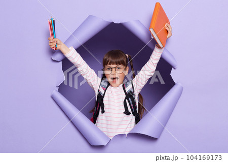 Horizontal shot of extremely happy little girl in a striped shirt with books and pencils, raised hands and screaming happily, wants to study, rejoicing. 104169173