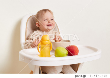 Indoor shot of satisfied little female girl wearing beige sweater sitting in high chair and eating, posing isolated over white background, having a snack with fruits and porridge. 104169256