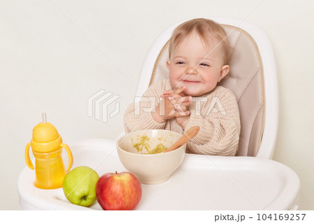 Portrait of cunning smiling little female girl wearing beige sweater sitting in high chair and eating vegetable puree, keeps hands together, isolated over white background. 104169257