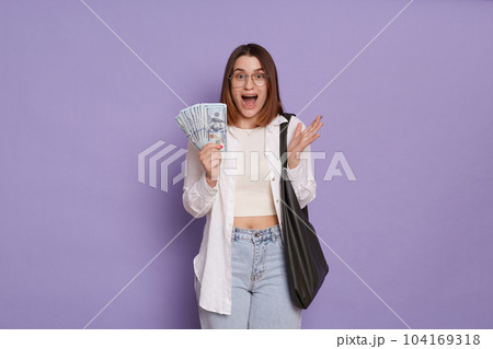 Portrait of amazed surprised overjoyed woman wearing stylish clothing holding bag and dollar banknotes, winning lottery, looks amazed, posing isolated over purple background 104169318