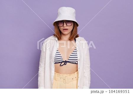 Portrait of confused surprised woman wearing panama, white shirt and swimming suit, looking at camera with dropped jaw, posing isolated over purple background. 104169351