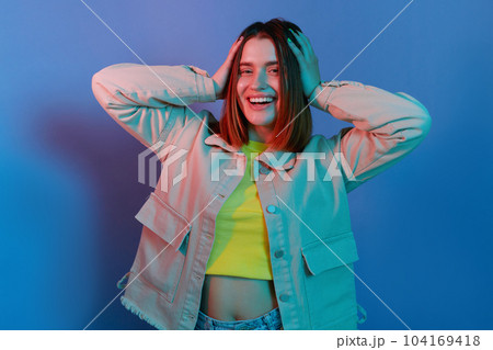 Indoor shot of smiling attractive, young girl wearing casual jacket standing isolated over blue neon light background, looking at camera, keeps hand on her head. 104169418