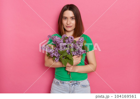 Horizontal shot of calm relaxed cute woman looks with pleasure at camera, wears green t shirt, holding lilac flowers, posing against pink background, charming female with bouquet. Horizontal shot of calm relaxed cute woman looks with pleasure at camera, wears green t shirt, holding lilac flowers, posing against pink background, charming female with bouquet. 104169561