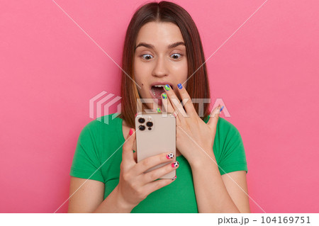 Portrait of excited shocked surprised woman wearing green t shirt, using smart phone, reading shocking breaking news in social networks, posing isolated over green background. 104169751