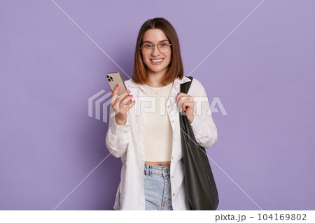 Image of adorable woman with black bag wearing white shirt and jeans posing isolated over purple background, using mobile phone, checking new app, looking smiling at camera. Image of adorable woman with black bag wearing white shirt and jeans posing isolated over purple background, using mobile phone, checking new app, looking smiling at camera. 104169802