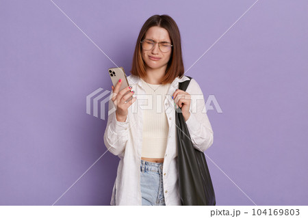 Indoor shot of sad depressed woman with black bag wearing white shirt and jeans posing isolated over purple background, standing with cell phone in hands and crying, having bad news. Indoor shot of sad depressed woman with black bag wearing white shirt and jeans posing isolated over purple background, standing with cell phone in hands and crying, having bad news. 104169803