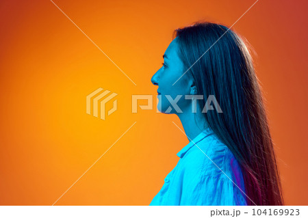 Side view portrait of young woman in casual clothes posing against gradient orange studio background in neon light. Profile 104169923