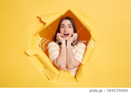 Portrait of surprised happy young dark haired female standing and looking at camera, expresses shock, keeps mouth opened and hands under chin, wearing striped t shirt, being in good mood. Portrait of surprised happy young dark haired female standing and looking at camera, expresses shock, keeps mouth opened and hands under chin, wearing striped t shirt, being in good mood. 104170000