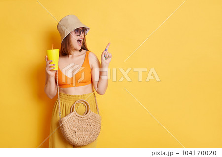 Indoor shot of astonished amazed woman in sunglasses wearing summer clothing posing isolated over yellow background, standing with fresh in hadns and pointing aside and advertising area. 104170020