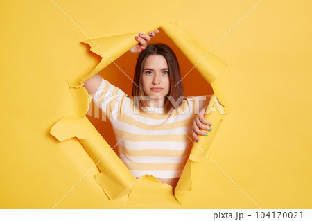 Horizontal shot of serious self-confident woman wearing striped T-shirt looking through hole in yellow paper wall, being strict and bossy, looks with confidence at camera. 104170021