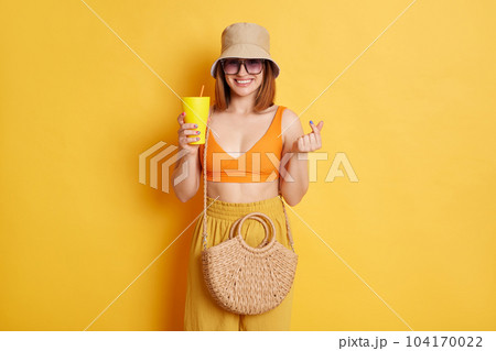 Image of lovely good looking woman in sunglasses wearing summer clothing posing isolated over yellow background, enjoying juice and showing korean love symbol. 104170022