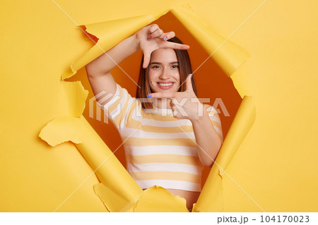 Portrait of smiling winsome woman photographer wearing striped T-shirt breaking through hole in yellow paper, looking through frame from fingers, making focus. Portrait of smiling winsome woman photographer wearing striped T-shirt breaking through hole in yellow paper, looking through frame from fingers, making focus. 104170023