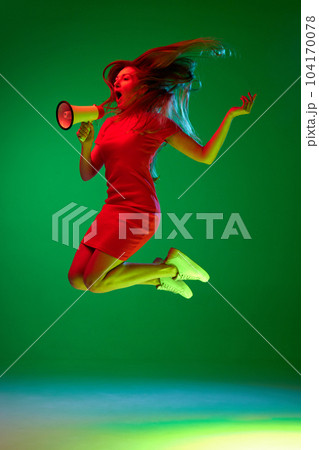Full-length portrait of young woman in beautiful red dress jumping and emotionally shouting in megaphone against green background in neon light 104170078