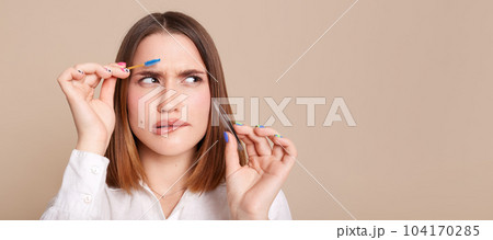 Indoor shot of puzzled confused woman in white shirt standing with eyebrow brush and tweezers in hands, looking away at copy space for advertisement or promotional text. Indoor shot of puzzled confused woman in white shirt standing with eyebrow brush and tweezers in hands, looking away at copy space for advertisement or promotional text. 104170285