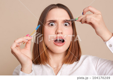 Portrait of shocked scared female in beauty salon holding eyebrow brush and tweezers, looking at camera with big eyes, being shocked after plucking eyebrow procedure. 104170289